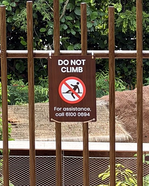 Do Not Climb sign on a timber barrier with wire mesh at an animal enclosure, with an assistance phone number